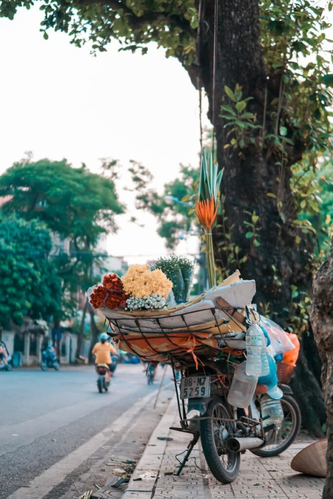 Ein Motorrad am Straßenrand in Asien mit einer übergroßen Ladefläche, auf der sich farbenfrohe Blumensträuße befinden. An der rechten Seite hängen mehrere leere Plastik-Wasserflaschen.